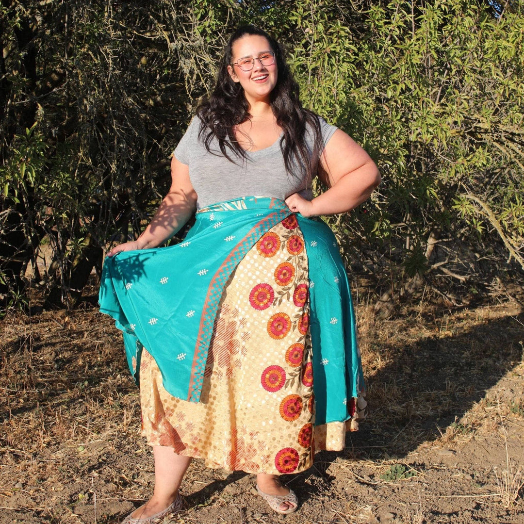 A Model standing in a field on a sunny summer day. She's wearing a goddess Sari Wrap Skirt, Size 14-32 in Maxi Length. The Under layer is gold with burnt orange accents with a teal top layer with cream colored leaves on it. She's paired the skirt with a light gray tee.