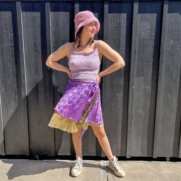 A model standing on a sidewalk wearing a petit 00-02 Sari wrap skirt in a mini length. The top layer of the skirt is a baby pink with purple floral patterns through out. the bottom later is a plain gold layer. She's paired the skirt with a baby pink ruffled tank top and pink bucket hat.
