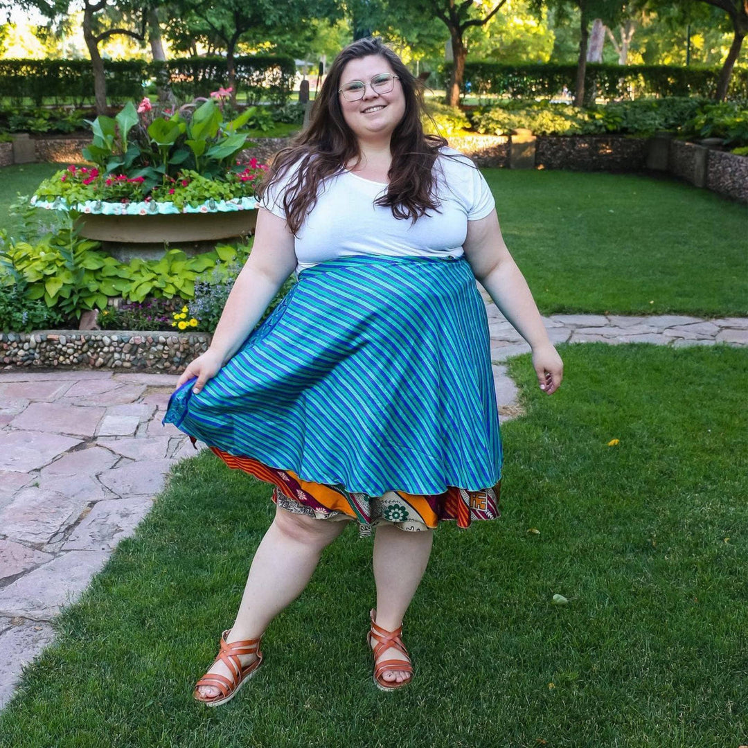 A model standing in a park on a spring day. She's wearing an Adjustable Size 14-32 Sari Wrap Skirt, mini length. The top layer is an aqua blue with stripes while the under layer is a burnt orange and red with floral designs. She's paired the skirt with a plain white tank top with a gladiator sandal.