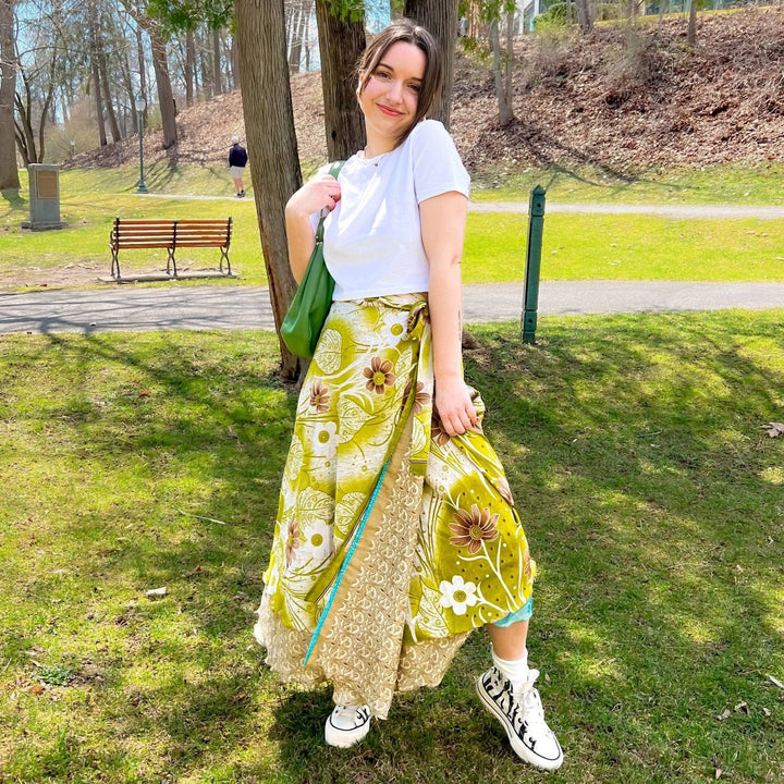 Woman in white top and floral yellow skirt in park. Green bag, white sneakers.