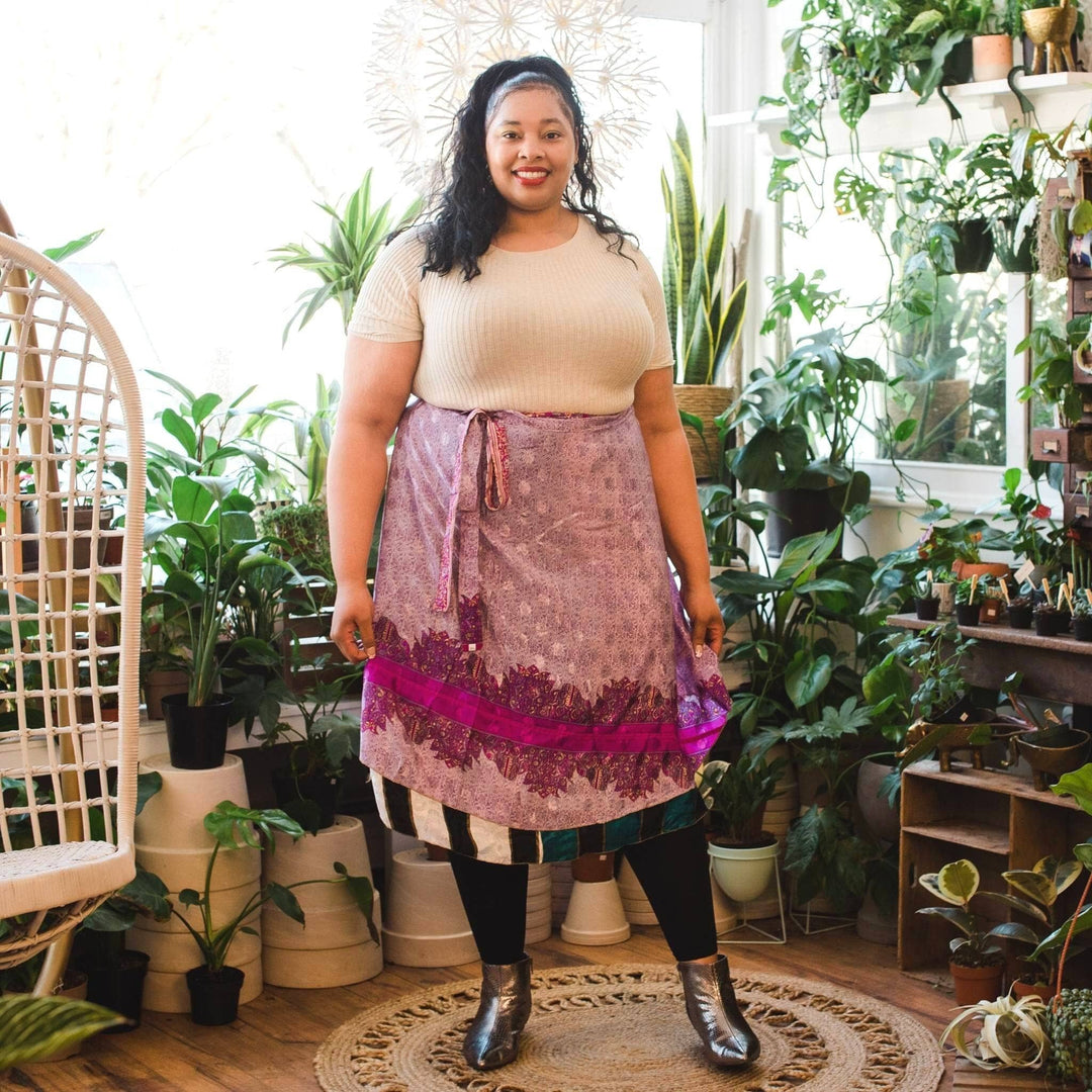 A model shopping on a floral shop. She's wearing a Goddess 14-32 Sari Wrap Skirt in a Tea length. The top layer is a baby pink with magenta accents. There's a beautiful floral pattern throughout. The under layer is a black and white stripe. She's paired this skirt with leggings, black booties and a cream colored top.