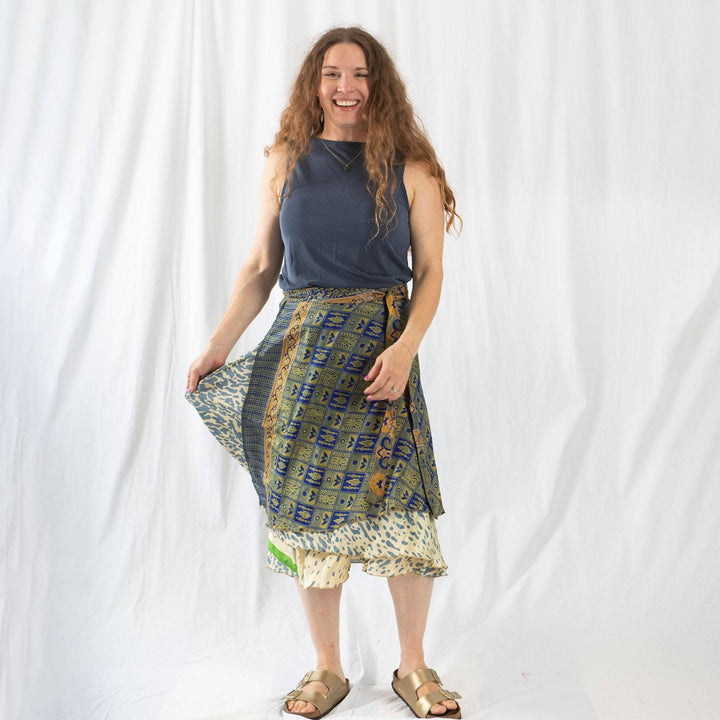 A model standing in front of a white wall. She's wearing a 02-12 Sari Wrap Skirt in a tea length. The skirt has navy, green and gold checkered squares all over. The lower layer is a cream base with navy and green specs. She's paired the skirt with a matching Navy tank top and strapy gold sandals.
