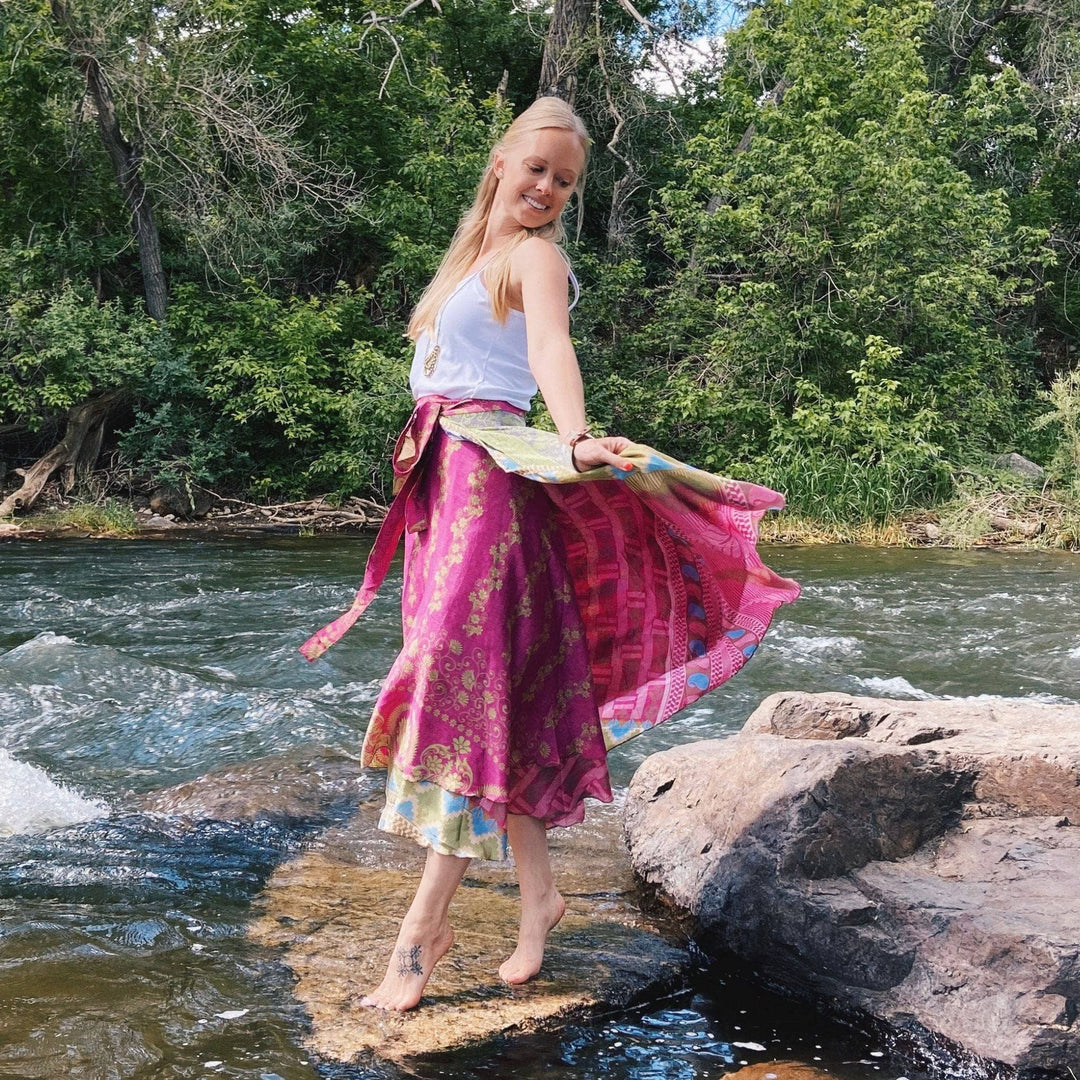 A model standing on rocks in a river. She's wearing a bright pink sari wrap skirt with gold accents. The Skirt is blowing in the breeze. She's paired it with a white tank top and bold gold jewlery