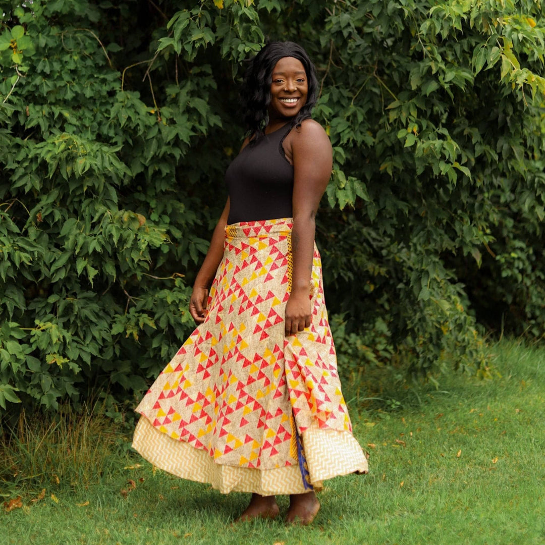 A Model standing in an open field wearing a 02-12 Sari Wrap Skirt in a Maxi Length. The top layer is a tan base with yarn and pink triangles all over. The lover layer is a light yellow and gold chevron pattern. She's paired the skirt with a simple black tank top.
