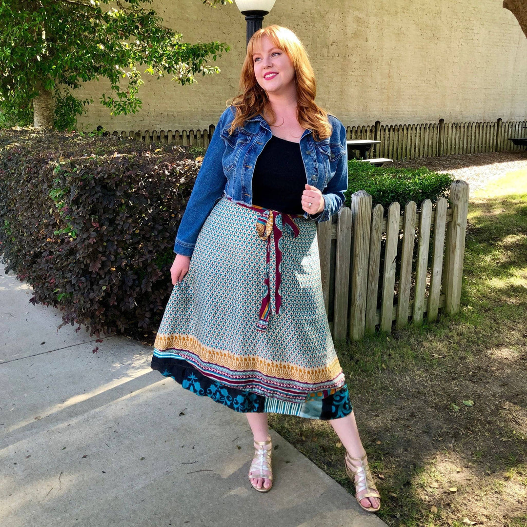 A model is standing in a park wearing a plus sized 08-20 Sari Wrap Skirt in a Tea Length. She's paired the skirt with a plain black tank top & short denim jacket and a gold gladiator sandal.