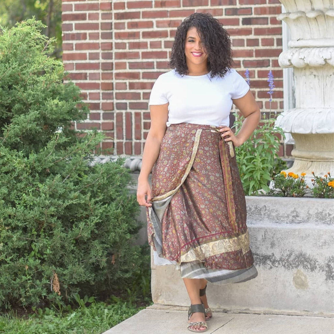 A model standing at a door step. She's wearing a Plus 08-20 Sari Wrap Skirt in a Maxi length. The top layer of the skirt is a rich brown with a cream colored floral pattern across it. The under layer is a white and grey stripe pattern. She's paired the skirt with a plain white tee and some gray open toed sandals.