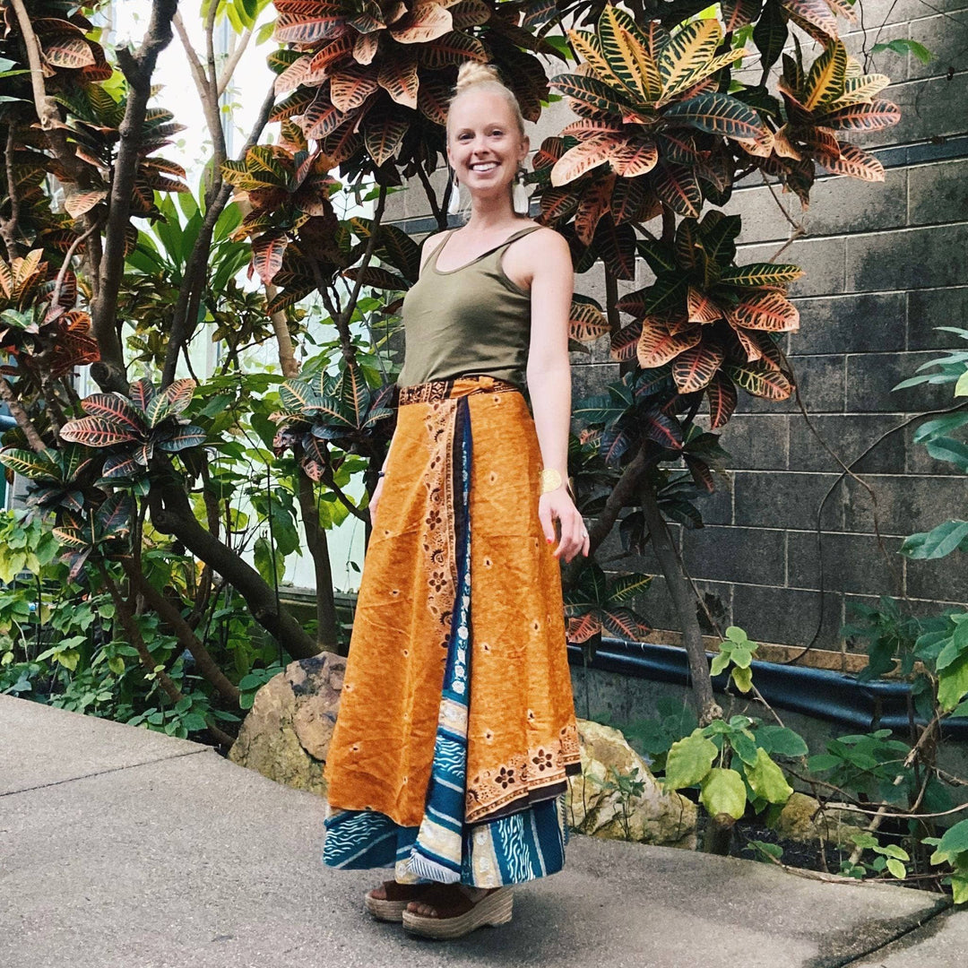 A model standing in a courtyard wearing a 02-12 Sari Wrap Skirt in a Maxi length. The top layer is a rich orange with dark blue floral around the lower edges. The under layer is a mix of teal and dark blue stripes. She's paired the skirt with a light green tank top and hunky heels.