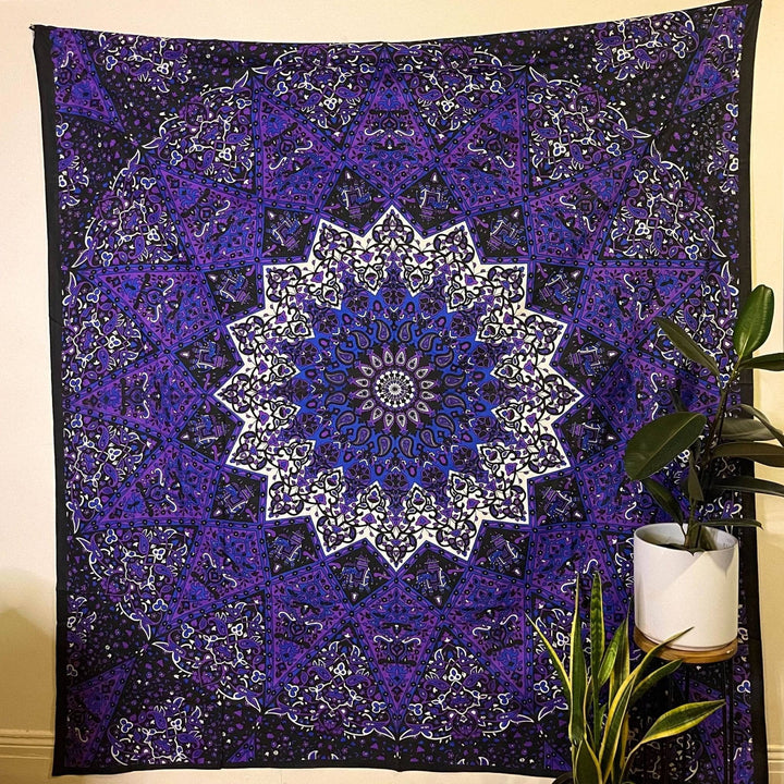 A Periwinkle Elephant Tapestry hanging on a cream colored wall. This tapestry is a black background with very intricate designs featuring elephants. This Tapestry has rich purples and whites  forming a big star pattern in the center.