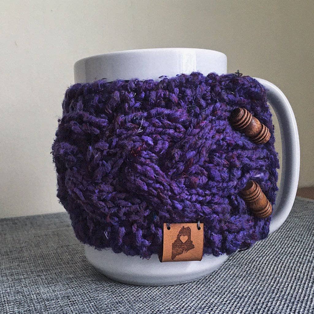 White mug with a purple knitted cozy with wooden buttons sitting on a gray fabric surface