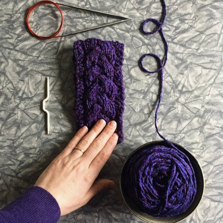 Purple knitted cozy with wooden buttons with purple yarn cake, circular knitting needles, and womans hand on a gray surface
