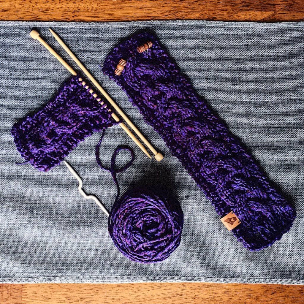 Purple knitted cozy with wooden buttons with purple yarn cake and knitting needles sitting on a gray fabric surface