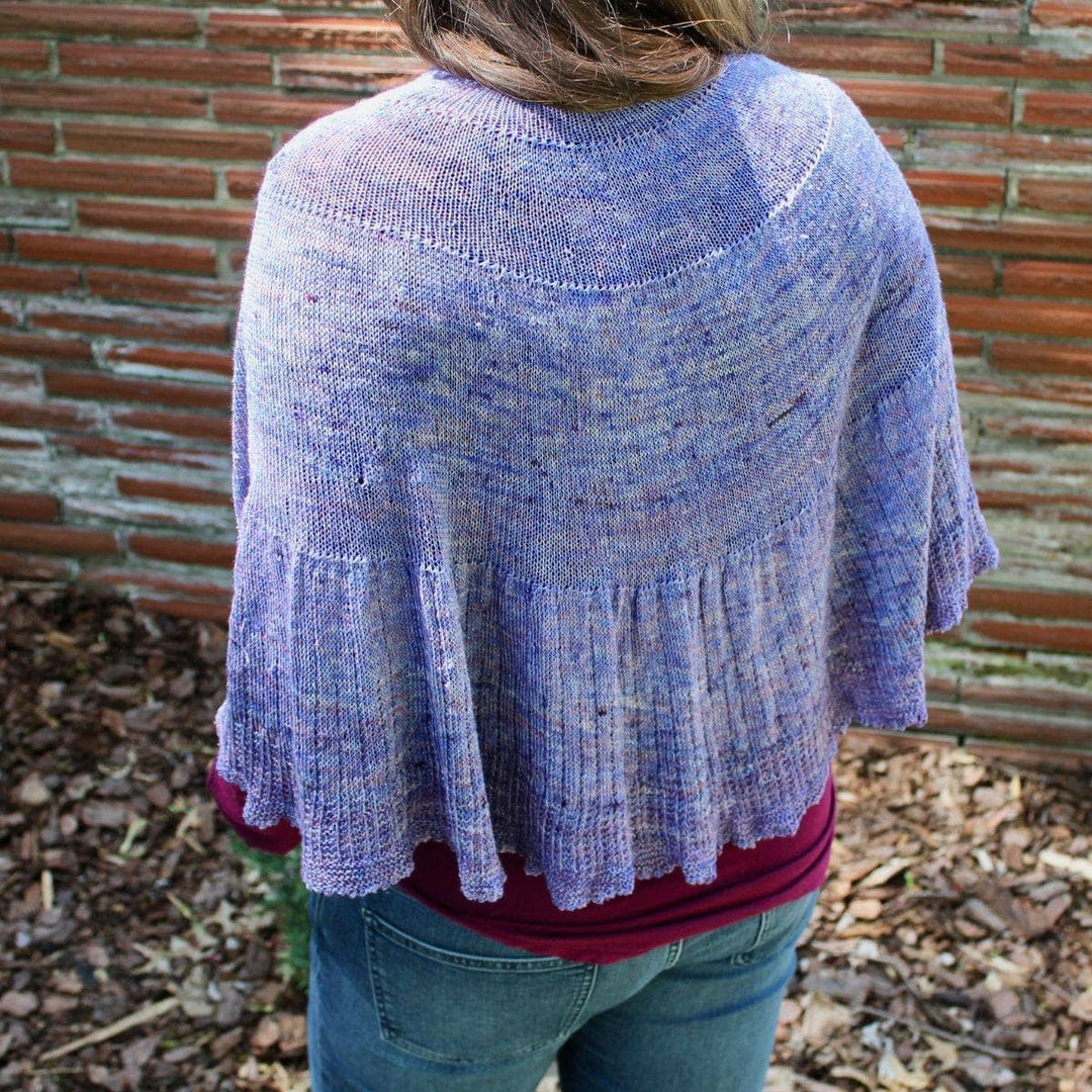 Back view of woman standing in front of a brick wall wearing a Calico Scallop Shawl in purple