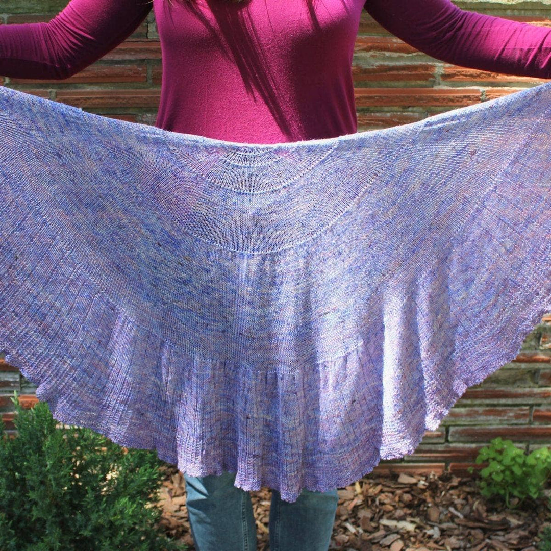 Woman standing in front of a brick wall holding up a Calico Scallop Shawl in purple