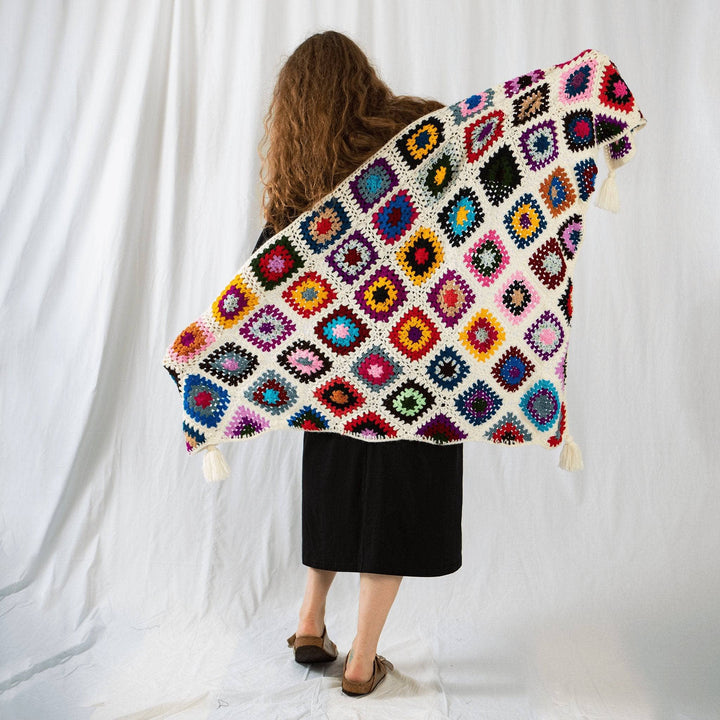 \Founder Nicole holding granny square shawl out behind her back while facing away from camera in front of a white background. Granny squares are three rounds of colors and a white border as well as white joins. 