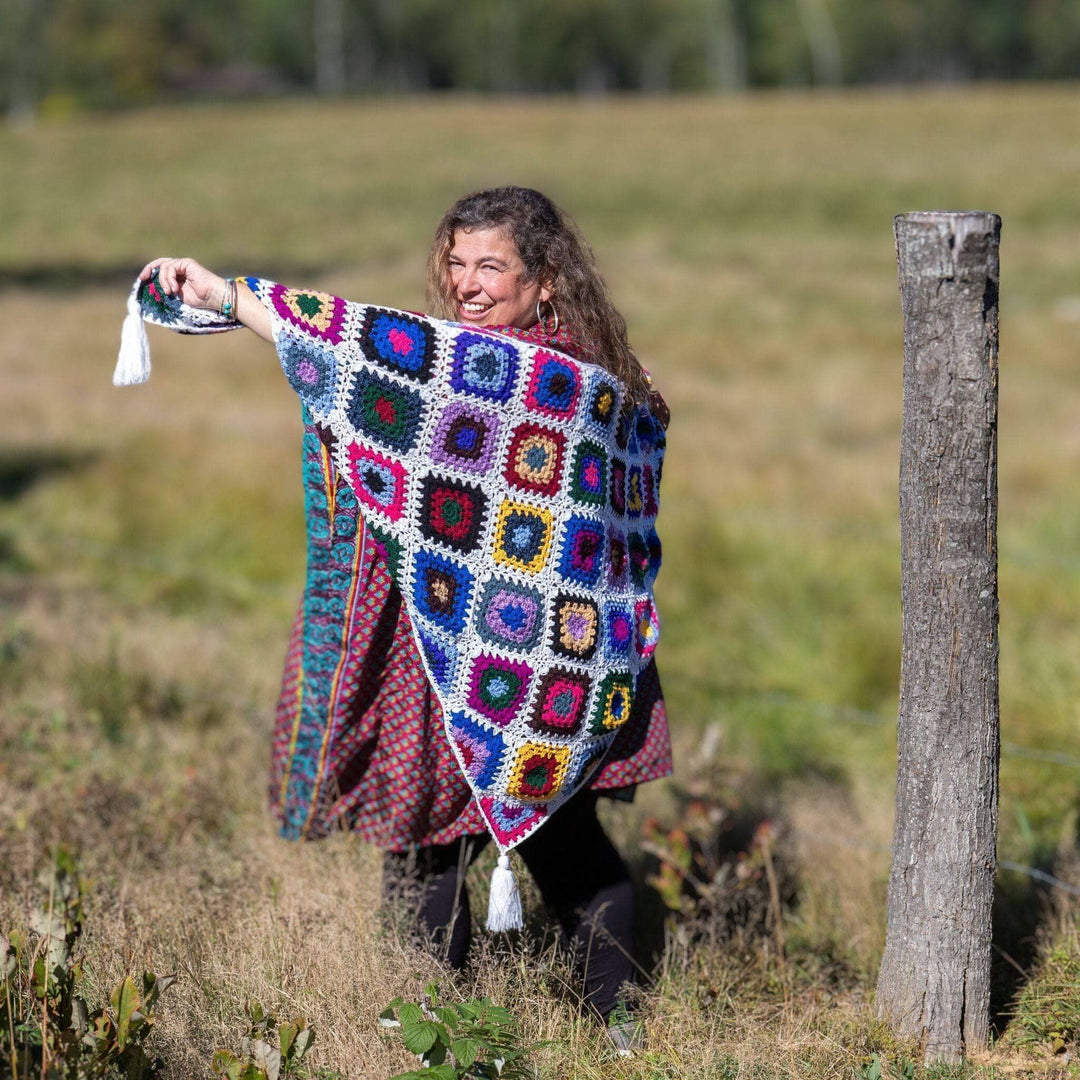 Model holding cape cod granny square shawl around themself while standing outside in a field with greenery in the background.