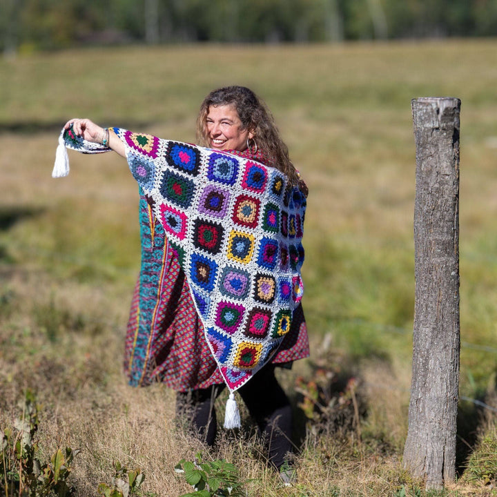Model holding cape cod granny square shawl around themself while standing outside in a field with greenery in the background.