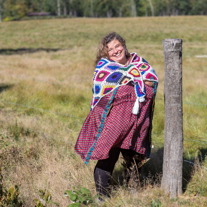 Model holding cape cod granny square shawl around themself while standing outside in a field with greenery in the background.