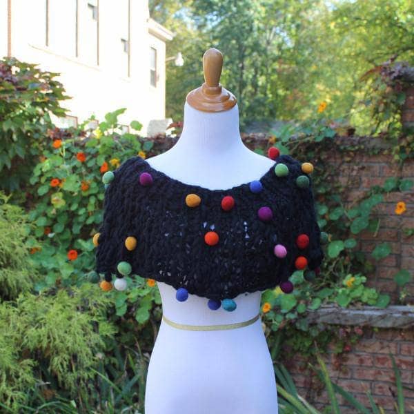 Mannequin wearing a Capowl Cowl in black with multicolored felt ball accents standing in front of greenery