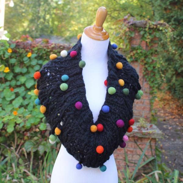 Mannequin wearing a Capowl Cowl in black with multicolored felt ball accents standing in front of greenery