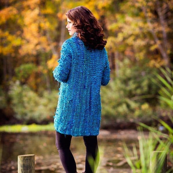 Back view of woman wearing the blue Catwalk Sweater with cowl neck and crossed front standing by greenery