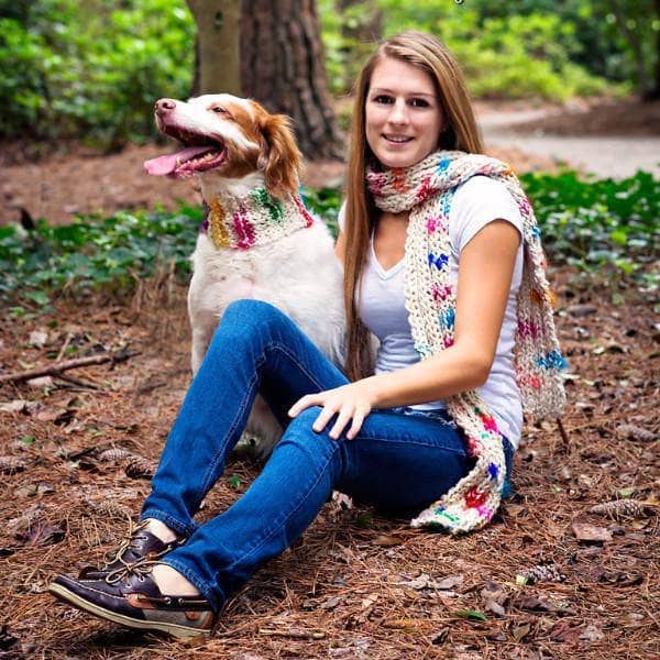 Woman walking her dog in the woods and wearing a multicolored knit Celebration scarf with a matching dog scarf