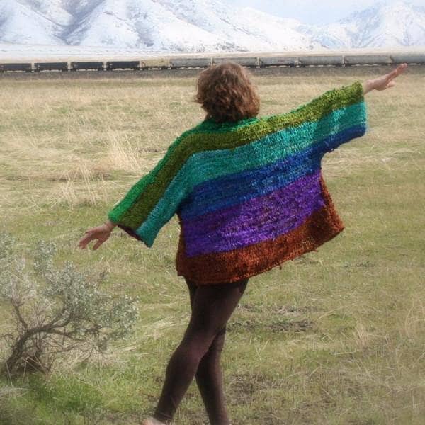 Back view of a woman wearing an oversized wide sleeve multicolor knitted jacket and standing in a grassy field