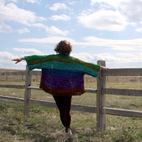 Back view of a woman wearing an oversized wide sleeve multicolor knitted jacket and standing in a grassy field