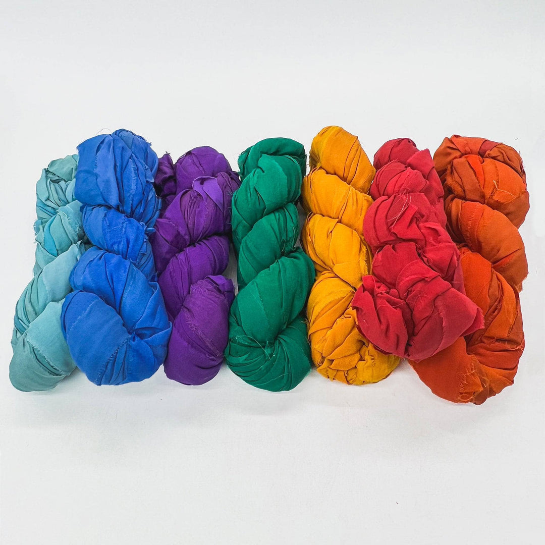 Rainbow chiffon ribbon yarn ombre pack in front of a white background. Left to right: light blue, true blue, violet, emerald green, golden yellow, red, orange spice.