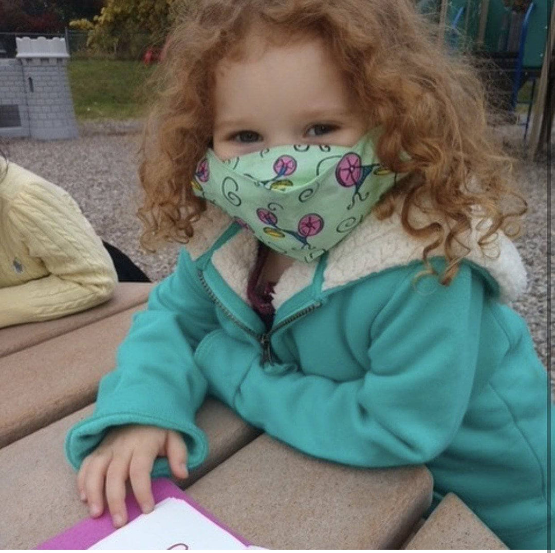 approx 3 year old child wearing green face mask outside at a picnic table with playground in the background.