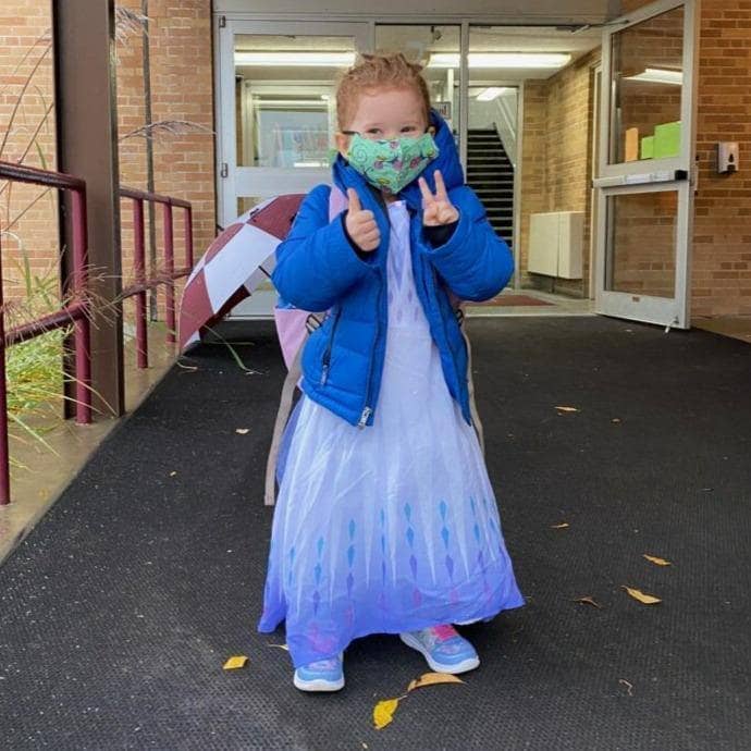 Approx 4 year old child wearing green face mask with blue dress giving thumbs up and peace sign. Brick school building with open glass door in the background.