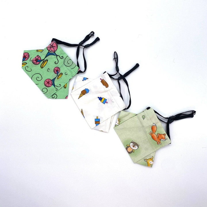 3 child size face masks in front of a white background. From left to right, green with pink and blue bicycles, white with multicolored ice creams, light green with multicolored forest animals.