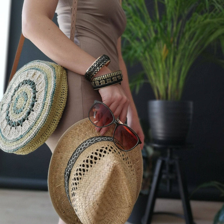 Woman wearing beige dress, circle banjo bag, straw fedora, and standing against a black wall with house plant