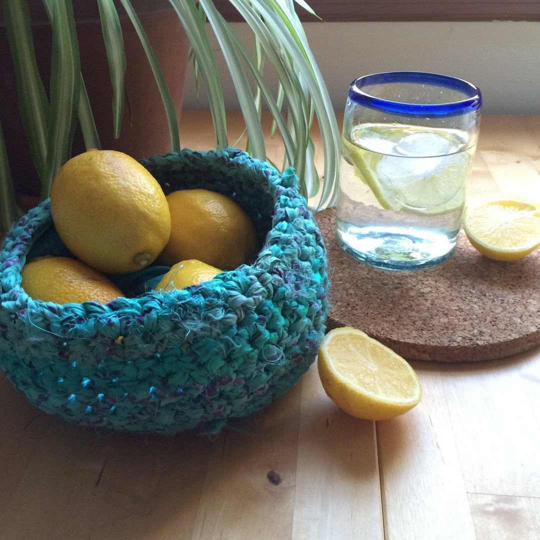 Blue knit bow filled with lemons next to a glass of water, sliced lemons, a cork coaster, and houseplant on a wooden surface