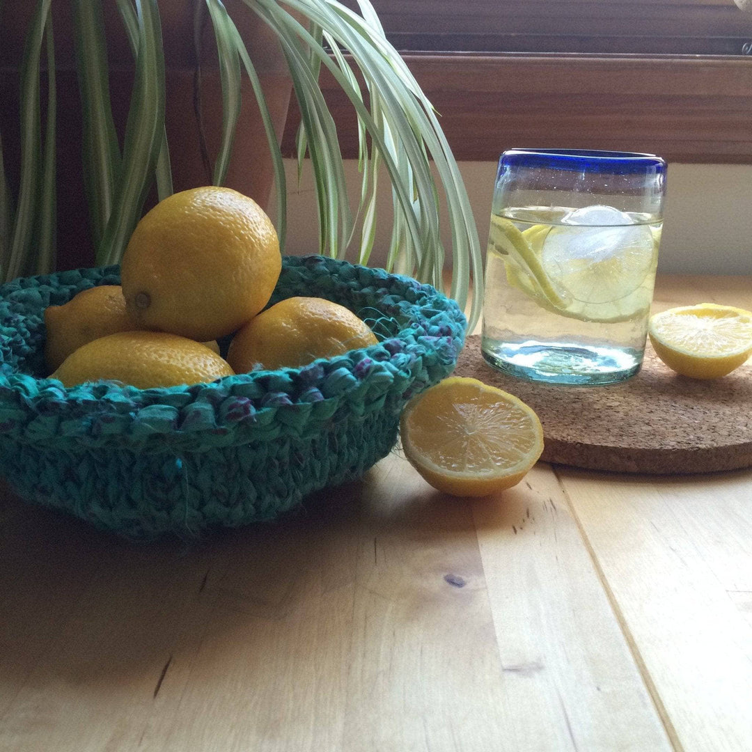 Blue knit bow filled with lemons next to a glass of water, sliced lemons, a cork coaster, and houseplant on a wooden surface