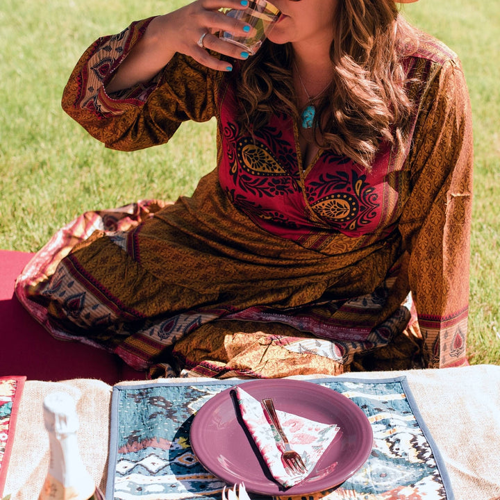 Model is sitting at a picnic with a table setting in front on them. A white cloth napkin sits on a plate with a fork on top. 
