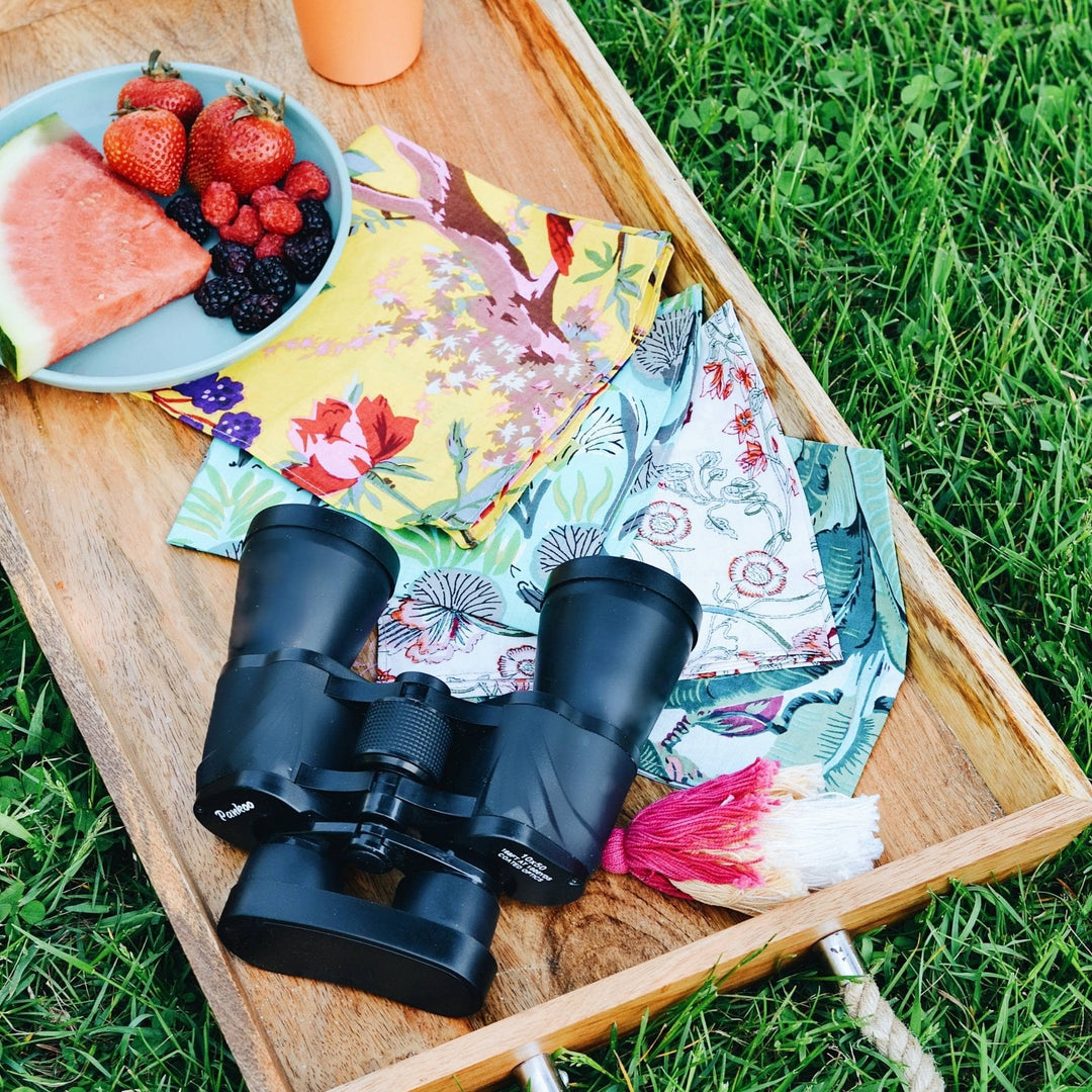 Assorted cloth napkins laid out on a wooden tray with binoculars and a bowl of fruit. 