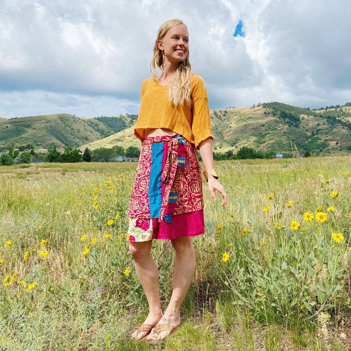 Blonde woman in yellow top, colorful skirt in meadow. Mountains and cloudy sky in background.