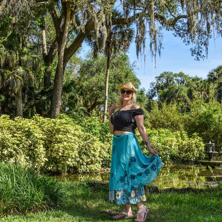 Woman in a garden wearing a blue skirt, black top, and straw hat, smiling.