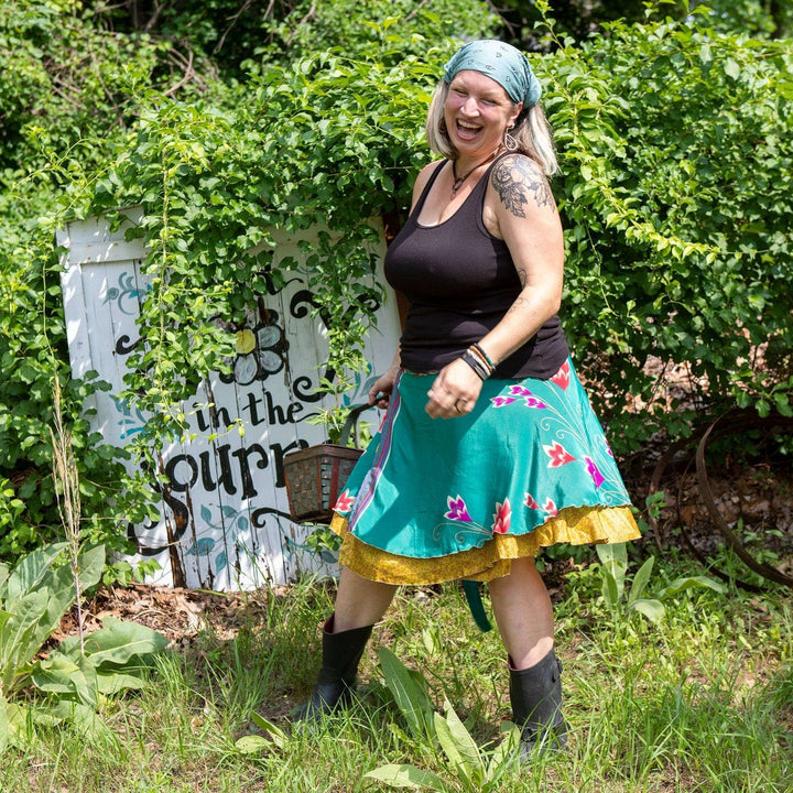 Woman laughing, wearing a colorful skirt and bandana, standing in a garden.