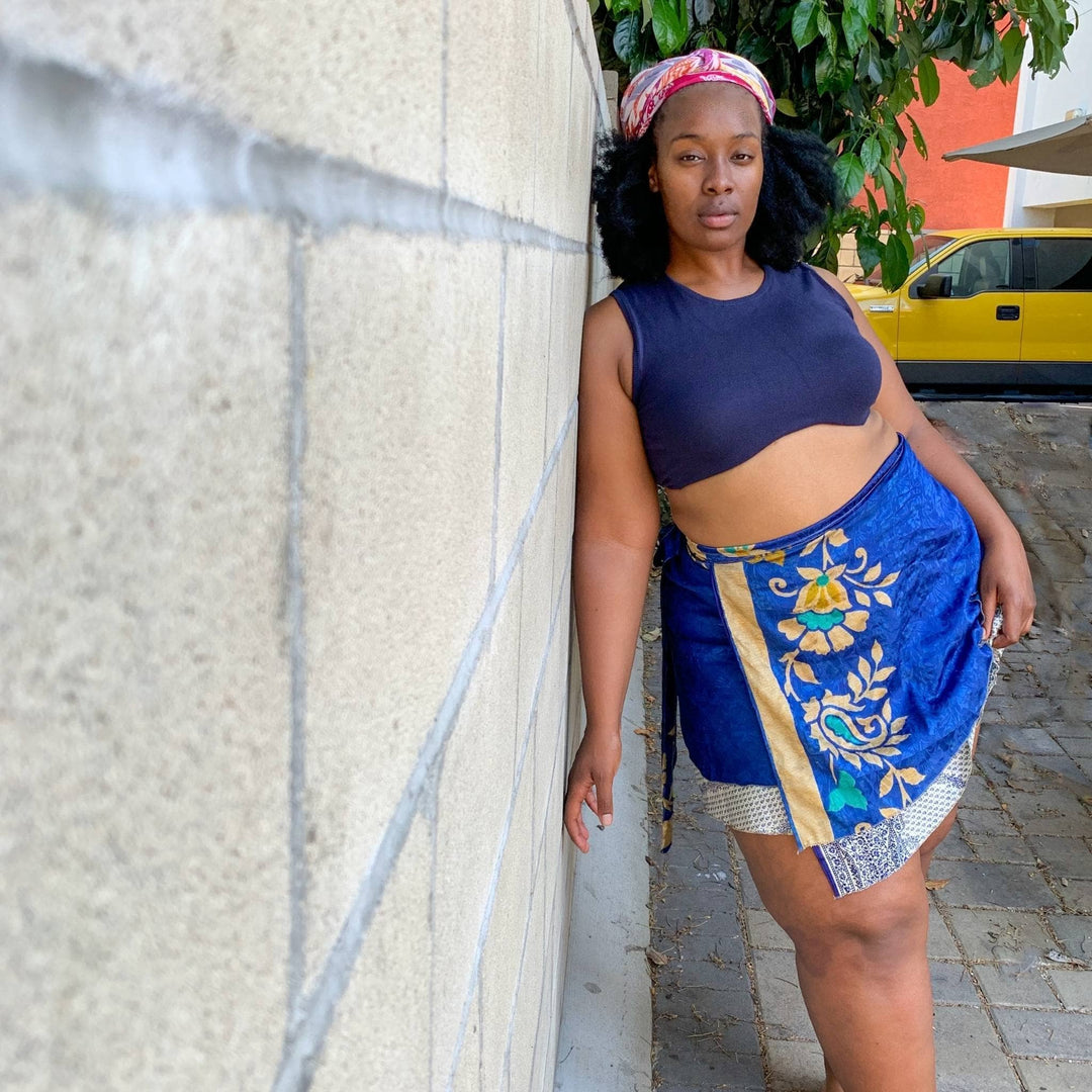 A woman leaning against a wall wearing a blue wrap skirt and black top.
