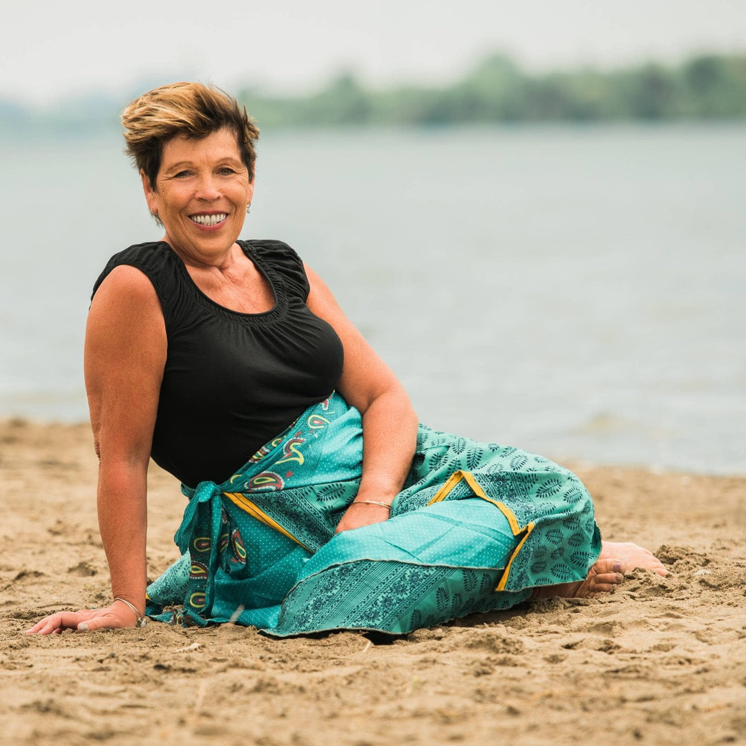 Mature woman with short hair, black top, teal patterned skirt, sitting on beach.