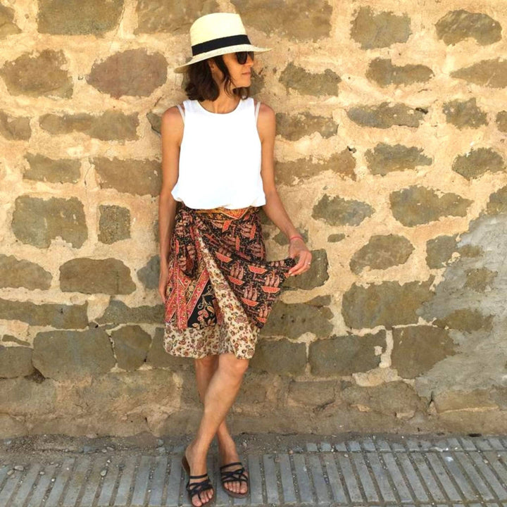 Woman in hat, white top, patterned skirt, posing against a stone wall.