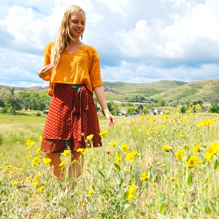 Woman in a field of yellow flowers, wearing a red wrap skirt and orange top.