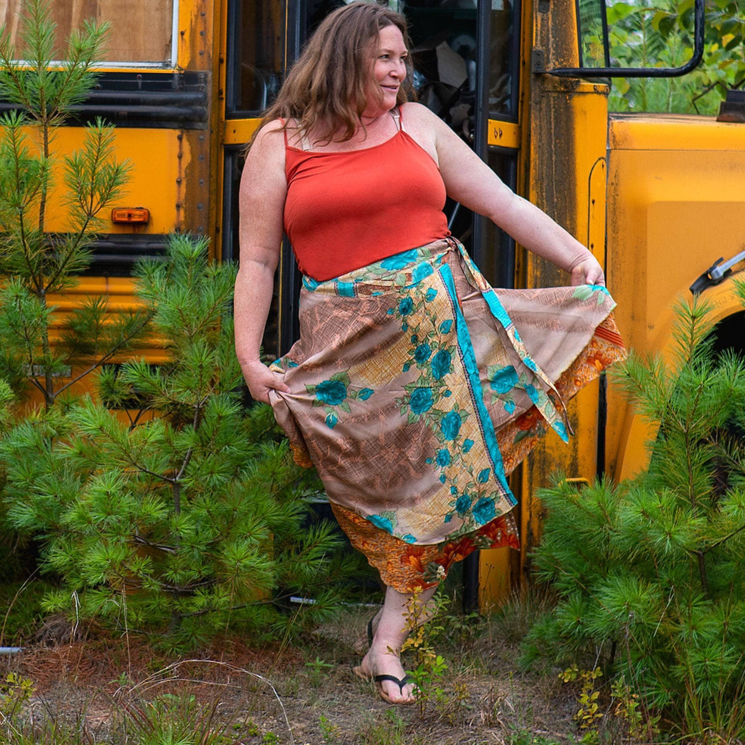 Woman in red top and floral skirt by yellow school bus. Surrounded by pine trees, smiling.
