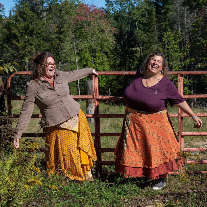 Two women laughing in a field, wearing colorful wrap skirts.