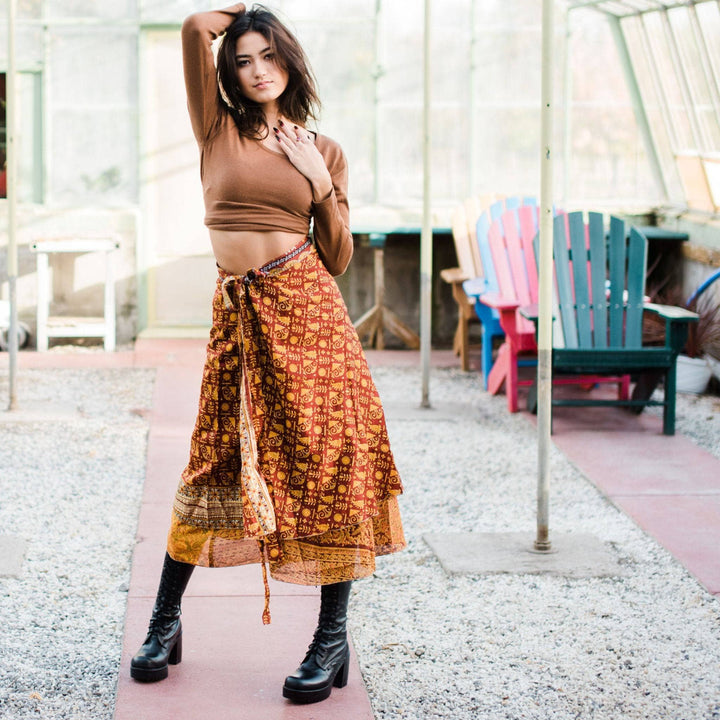 Woman in brown crop top, patterned skirt in greenhouse. Colorful chairs behind.