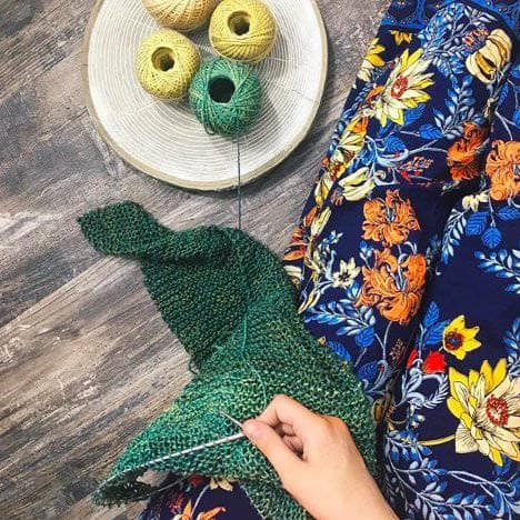 bird's-eye-view of woman wearing blue floral pants and knitting a shawl with green and yellow yarn cakes while sitting on a wooden surface