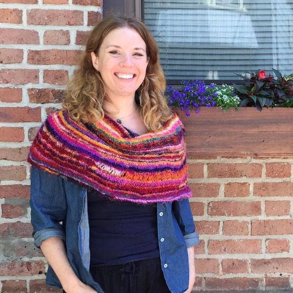 Woman wearing a denim shirt and multicolored striped knit shawl and standing in front of a brick wall