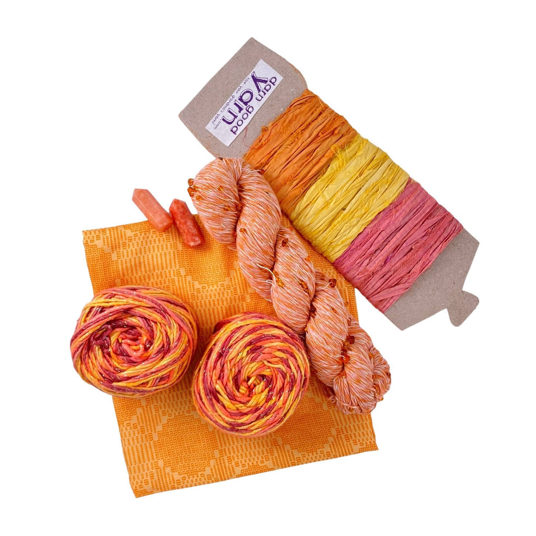 Orange Calcite energy bundle with all items showing in front of a white background. 2 orange calcite crystals, 2 skeins sparkle worsted weight silk, 1 skeins chakra beaded cotton yarn, 3 color sari silk ribbon sample card, and coordinating furoshiki.