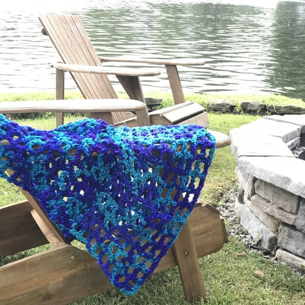 Blue and turquoise crochet blanket draped over adirondack chairs near a lake and a firepit